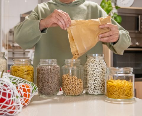 Person filling reusable glass jars from a paper bag with various legumes and pasta, promoting zero waste, plastic-free, and sustainable living practices in the kitchen pantry